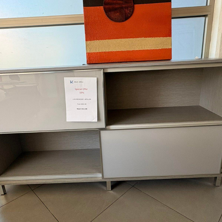 A light gray, modern bookcase with shelving and a glossy door sits beneath a colorful, abstract artwork near a window.