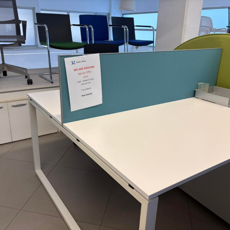 A white office desk with a teal divider stands in a showroom with office chairs displayed in the background.
