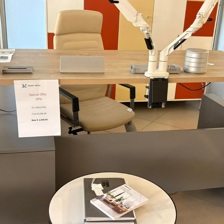 A beige office chair and a wooden desk with a white monitor arm, and a round table with books are shown.