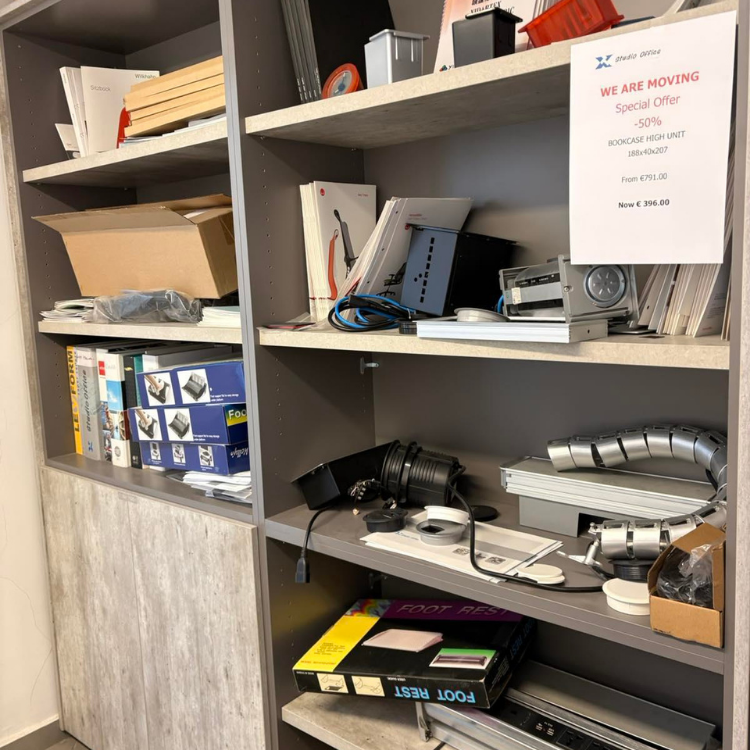 A tall gray bookcase with multiple shelves filled with various office supplies and equipment.