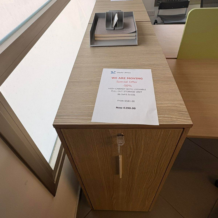 A wooden cabinet with a key in the lock is placed under a desk, with a flyer on top and office supplies.