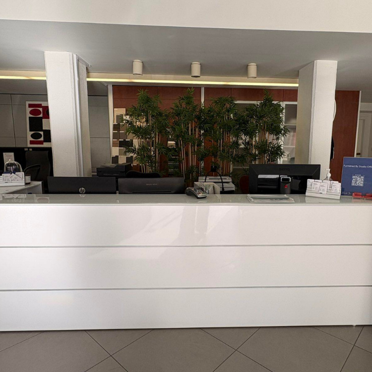 A white reception desk with two computers, a printer, and bamboo plants behind it in an office setting.
