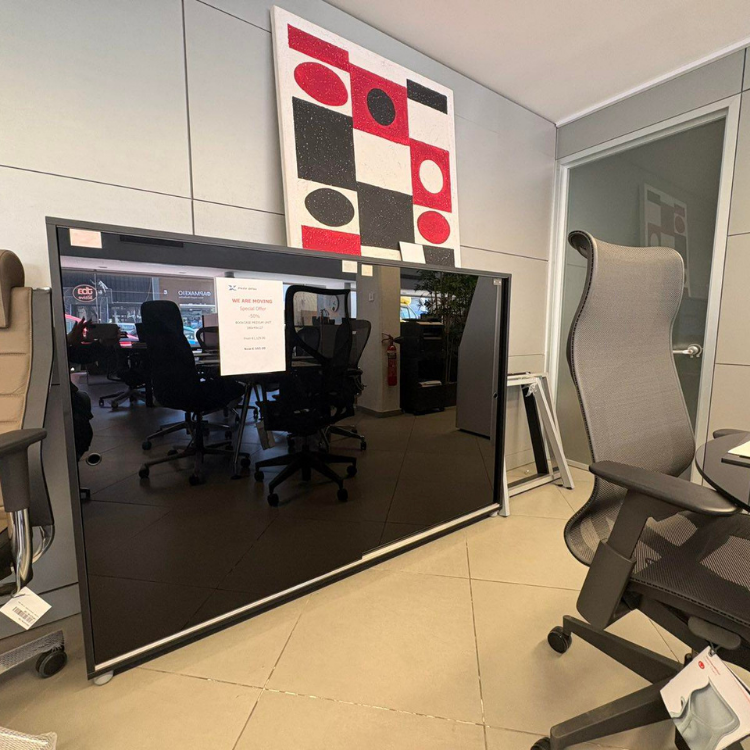 Современное офисное помещение со стульями и большой стеклянной доской в черной рамке, отражающей интерьер и абстрактную картину на стене.