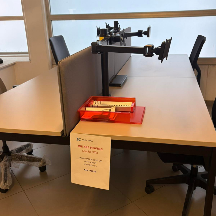A workstation desk with monitor arms, office supplies in a red container, and a promotional sign.