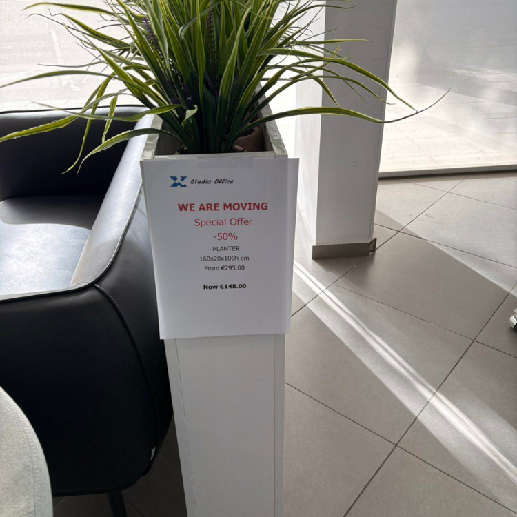 A tall, white planter with green plants sits next to a black couch, with a promotional sign attached.