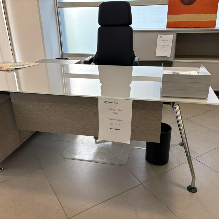 A desk with a light wood finish, a white top, and chrome legs stands in an office with a black chair.