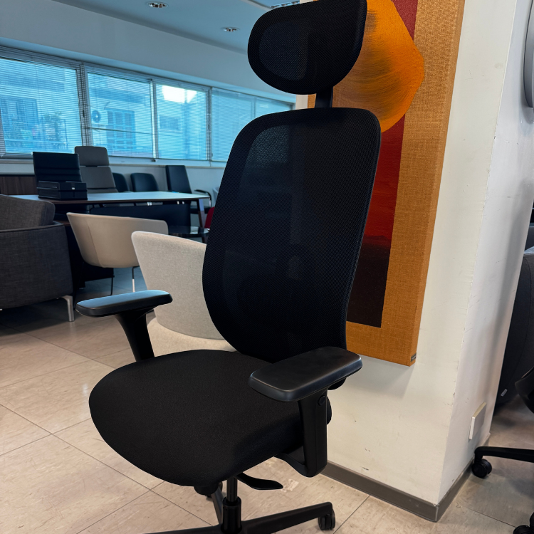 A black office chair with a mesh back and headrest sits in a showroom with other furniture visible in the background.