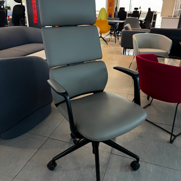 A gray office chair with a headrest and black armrests sits on a tiled floor in a showroom.