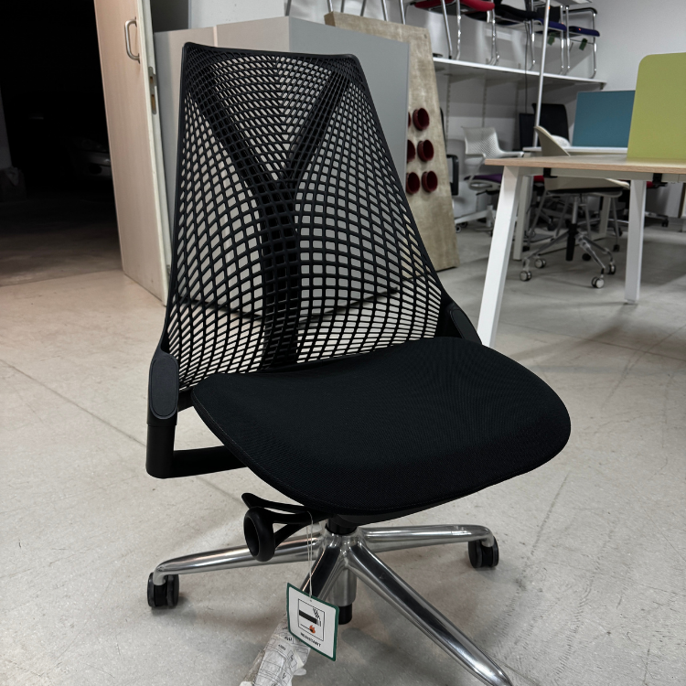 A black office chair with a mesh back and a silver base sits in a room with other office furniture.