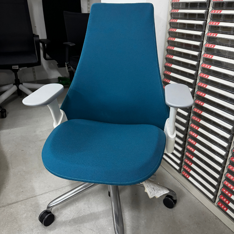 A blue office chair with a high back, gray armrests, and a chrome base is shown in a warehouse setting.