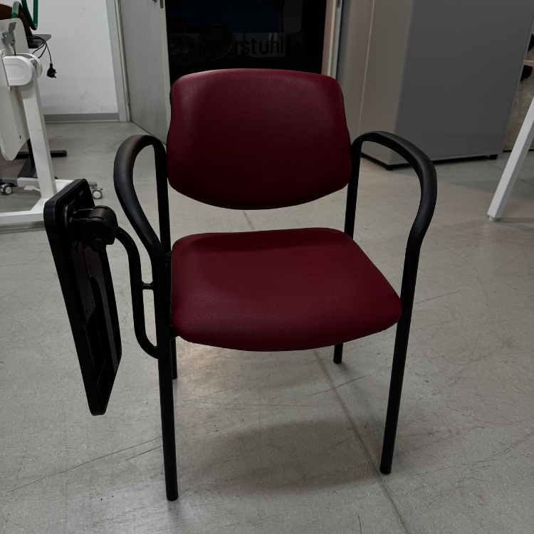 A maroon upholstered chair with black metal frame and attached black writing tablet is pictured indoors.