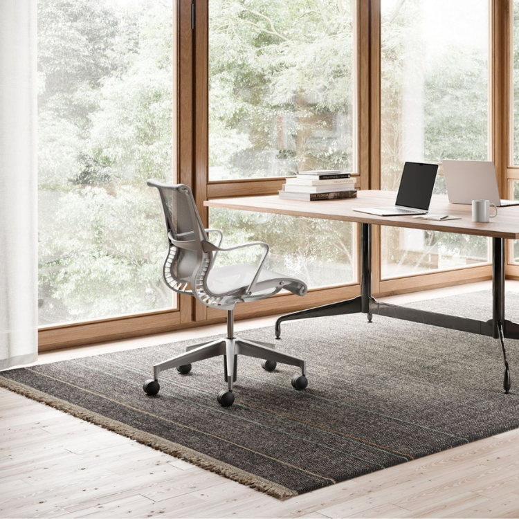 An ergonomic silver task chair is positioned at a wooden workstation in a room with large windows.