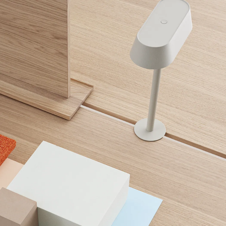 A minimalist light grey table lamp stands on a light-colored wooden surface next to geometric blocks and a wooden panel.