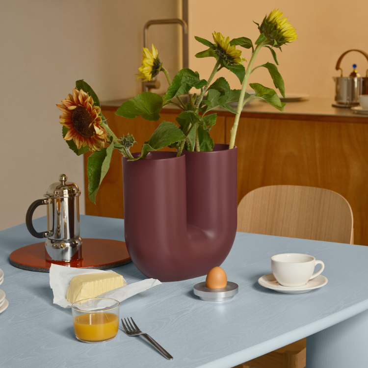 A maroon U-shaped ceramic vase holding sunflowers sits on a light blue table set for breakfast.